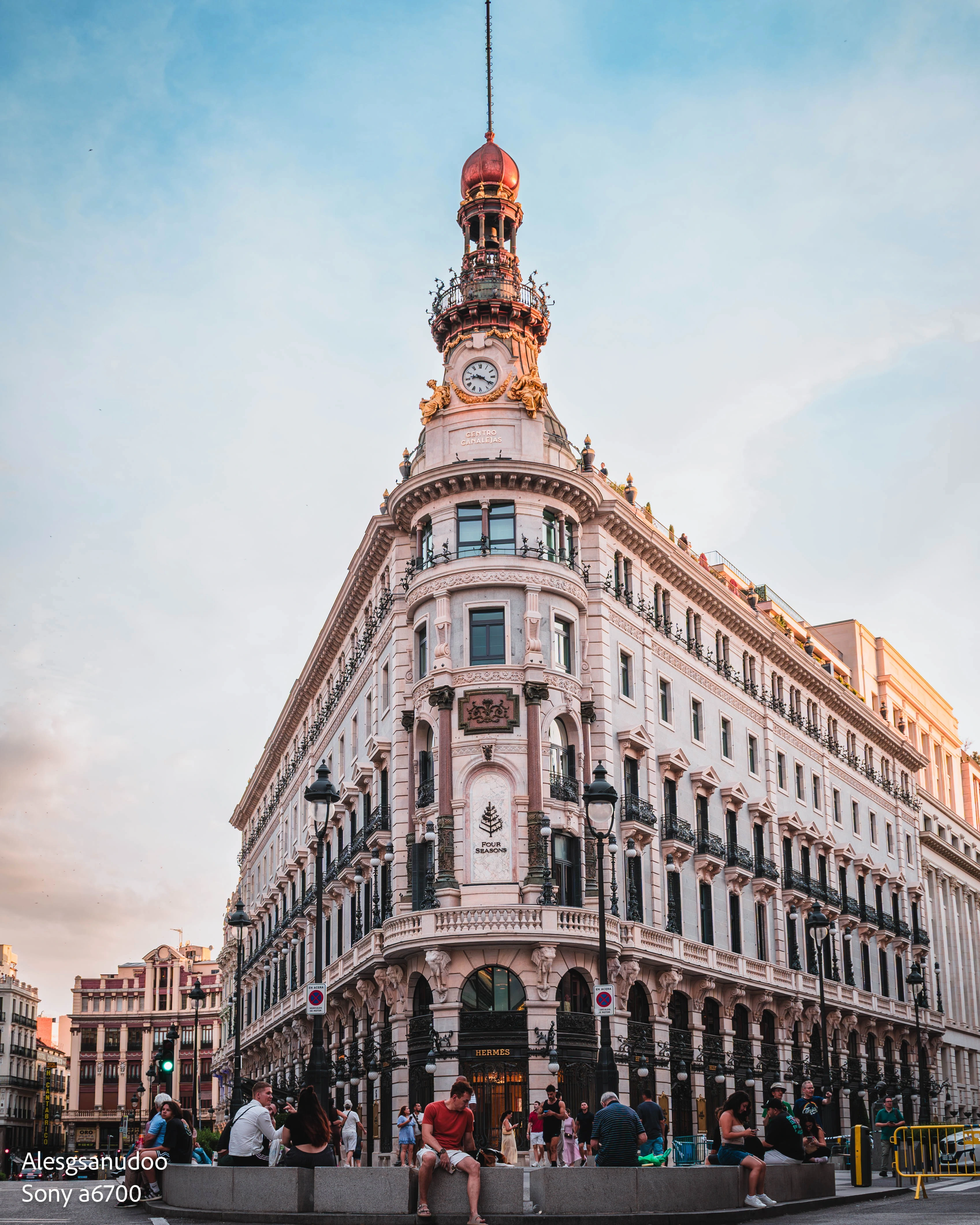 Edited street photo of an ornate building with warm highlights, soft blue sky, and people in the foreground.